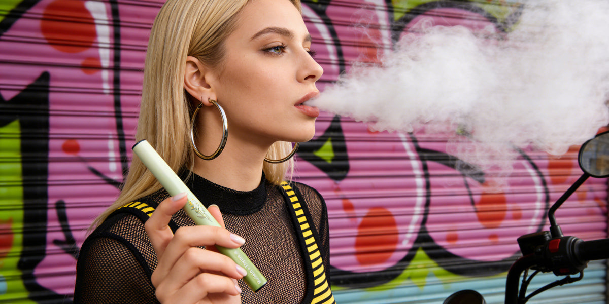 Blonde woman with large hoop earrings holding a green HOLYMOLY Mint herbal diffuser while exhaling white aromatic vapor in front of a colorful graffiti wall